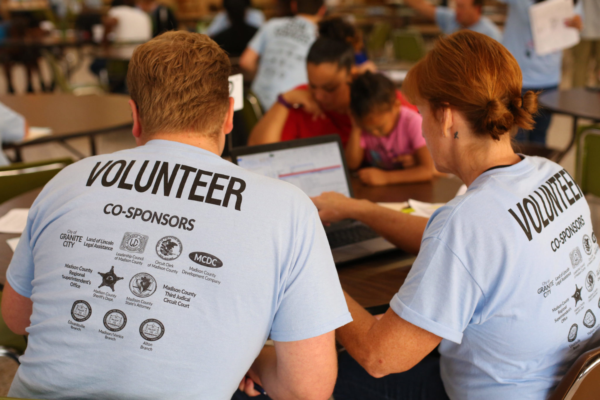 Two people wearing light blue shirts that read "volunteer" look at a computer